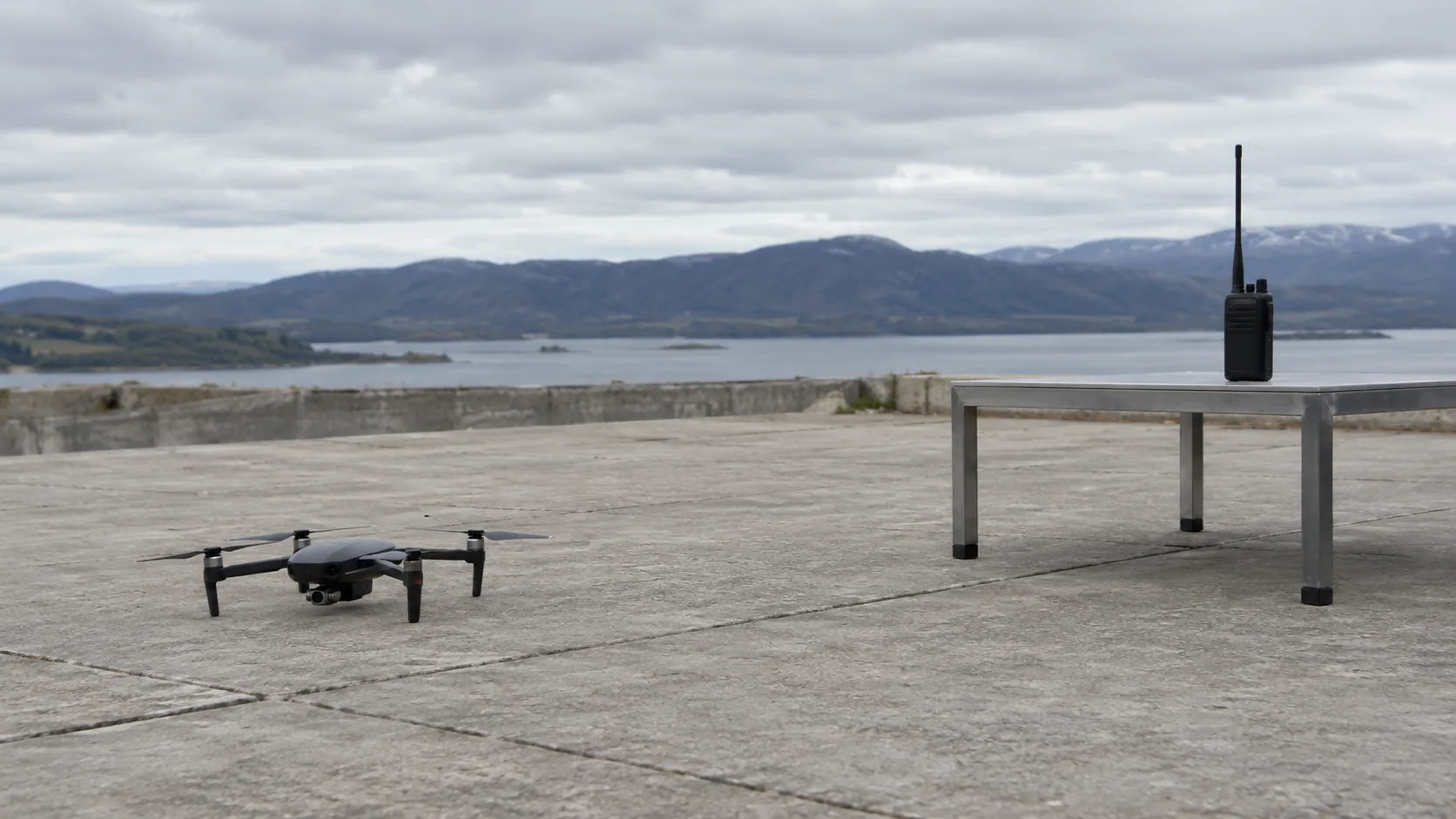 Unmarked quadcopter on a concrete pad, with a handheld radio standing on a metal table